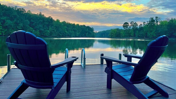 Two Adirondack chairs on a wooden deck overlooking a calm lake with a forested shoreline. - Lakefront Treehouse Retreat Minutes from Clemson University! (Seneca)