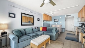 A compact living space with a sofa, a small wooden bench, a kitchenette, and a ceiling fan. - Oceanfront Studio-Sleeps4-Linens Incl-WiFi-Kitchen (Ocean City)