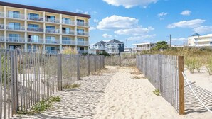 A sandy pathway leading to a multi-story building with balconies, surrounded by a wooden fence. - Oceanfront Studio-Sleeps4-Linens Incl-WiFi-Kitchen (Ocean City)