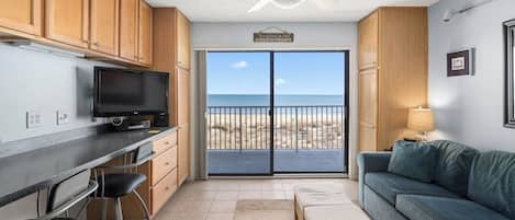 A compact living space with a kitchen, a dining area, a sofa, and a balcony with a view of the beach.