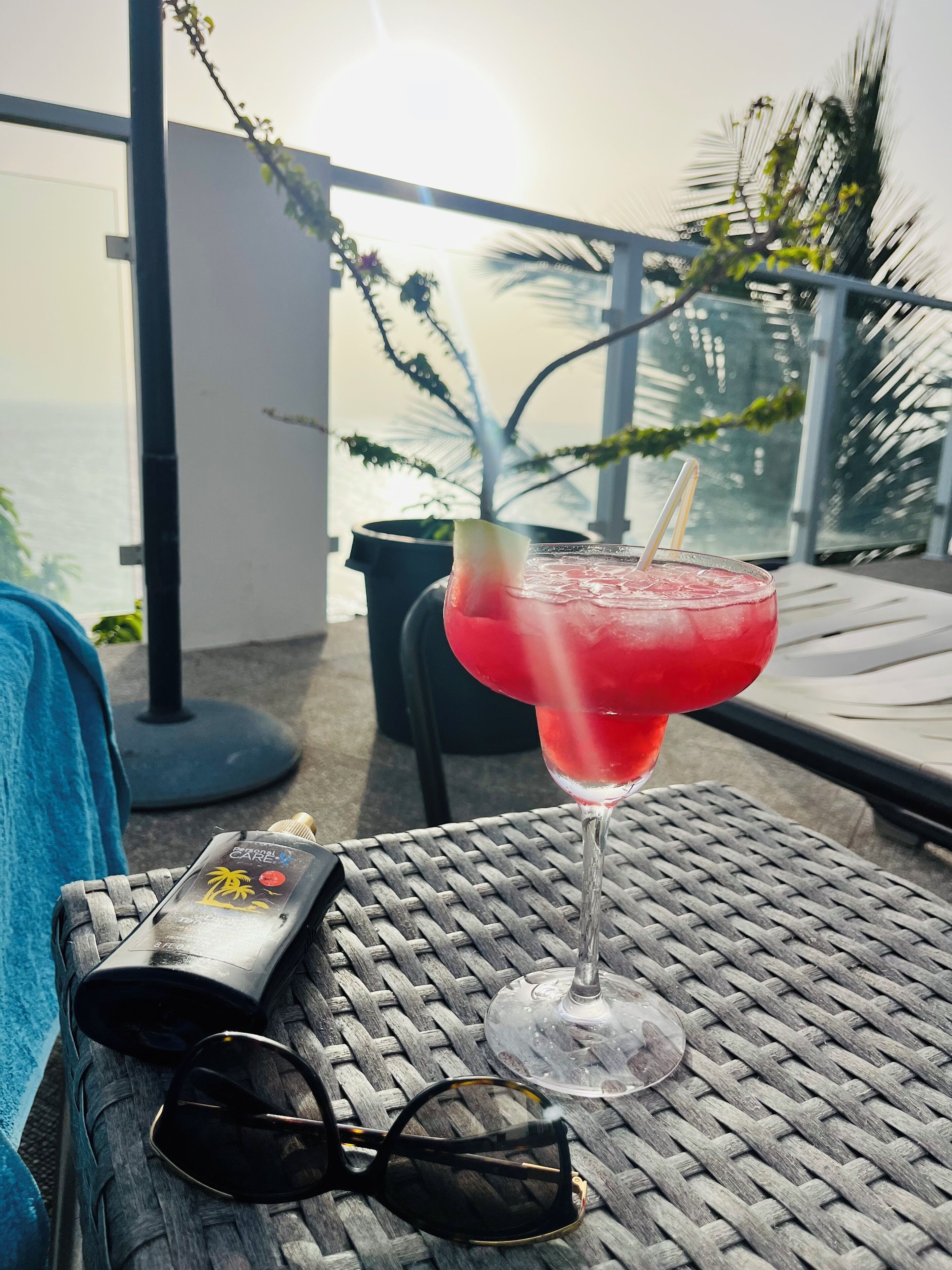 Una mesa con una bebida, gafas de sol y una botella de protector solar en un balcón con vista al océano.