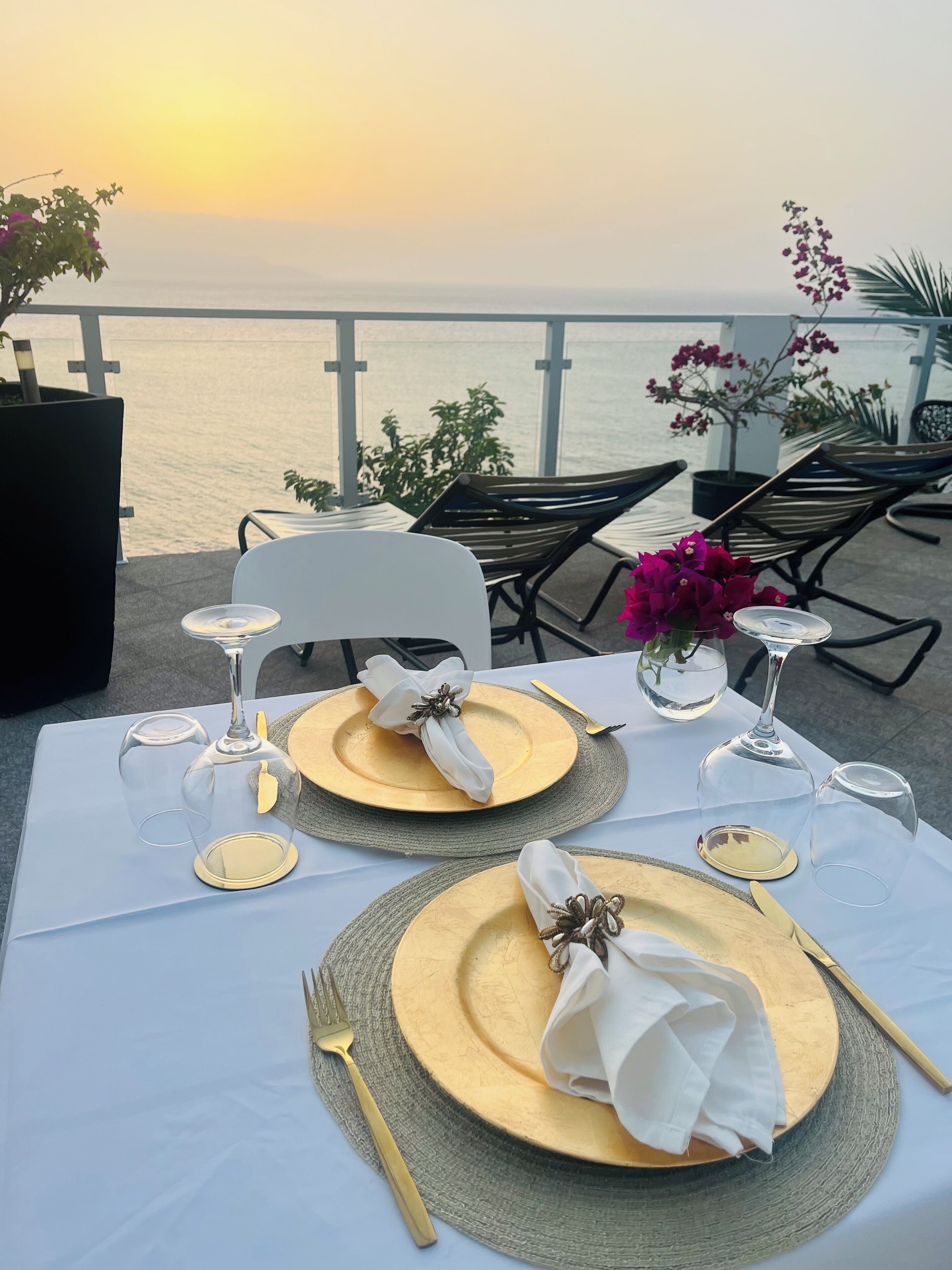 A table set for a meal with gold plates, white napkins, and wine glasses, overlooking a body of water.