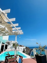 Zona junto a la piscina con una pérgola, sillas de descanso y vista al océano.