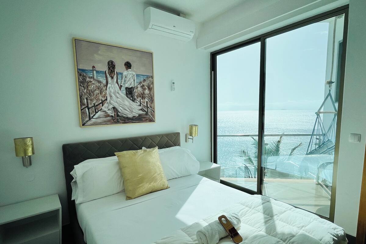 Une chambre d’hôtel avec un grand lit, un tableau représentant un couple sur un balcon et une vue sur la mer.