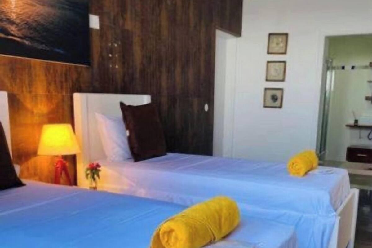 Une chambre d’hôtel avec deux lits, chacun recouvert d’une couverture bleue et agrémenté d’une serviette jaune. On y trouve une tête de lit en bois, une lampe et un tableau accroché au mur.