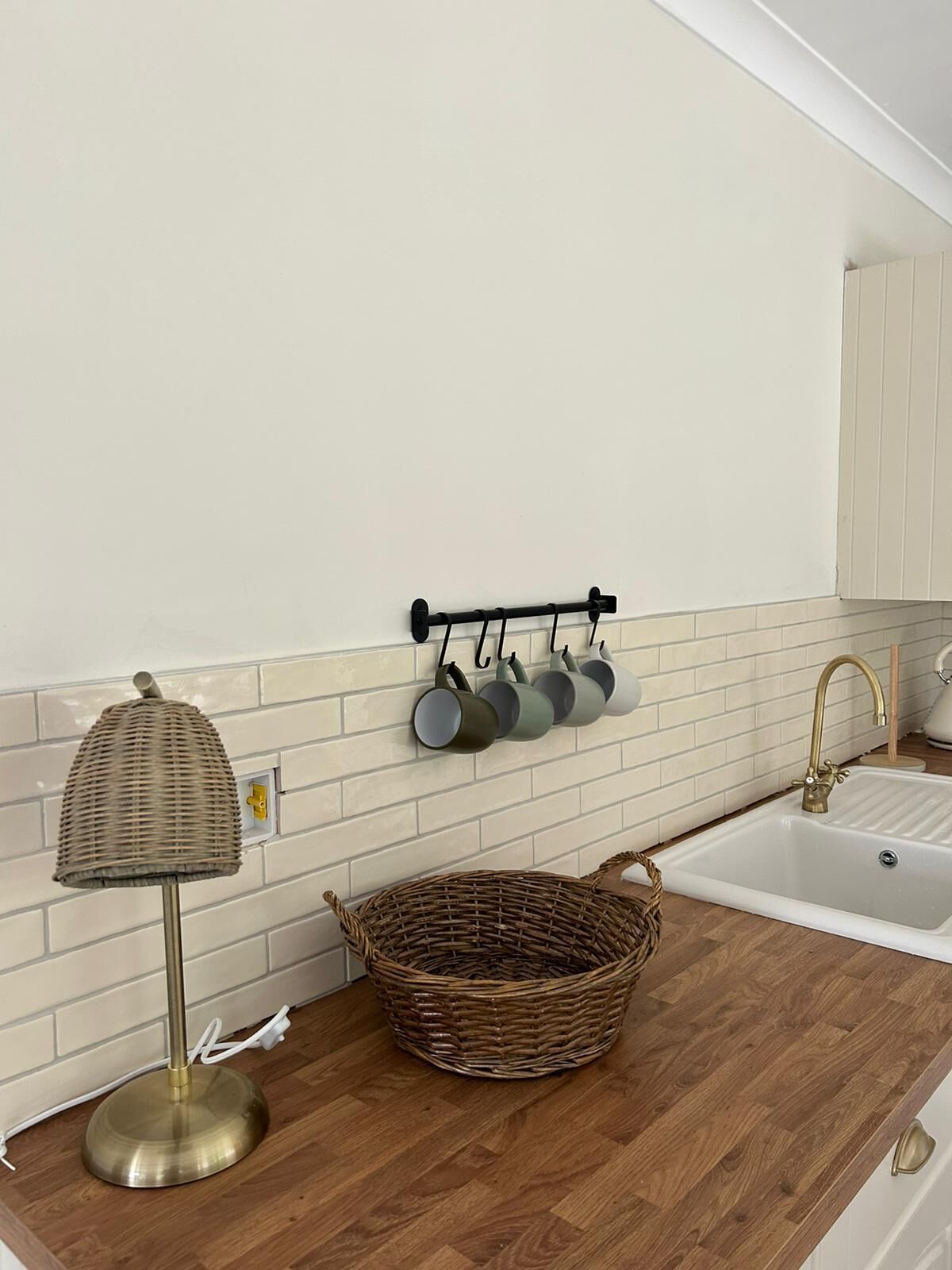 Una cocina con salpicadero de azulejos blancos, encimera de madera, un colgador para ollas con varias ollas, una cesta de mimbre y una lámpara de pie de latón.