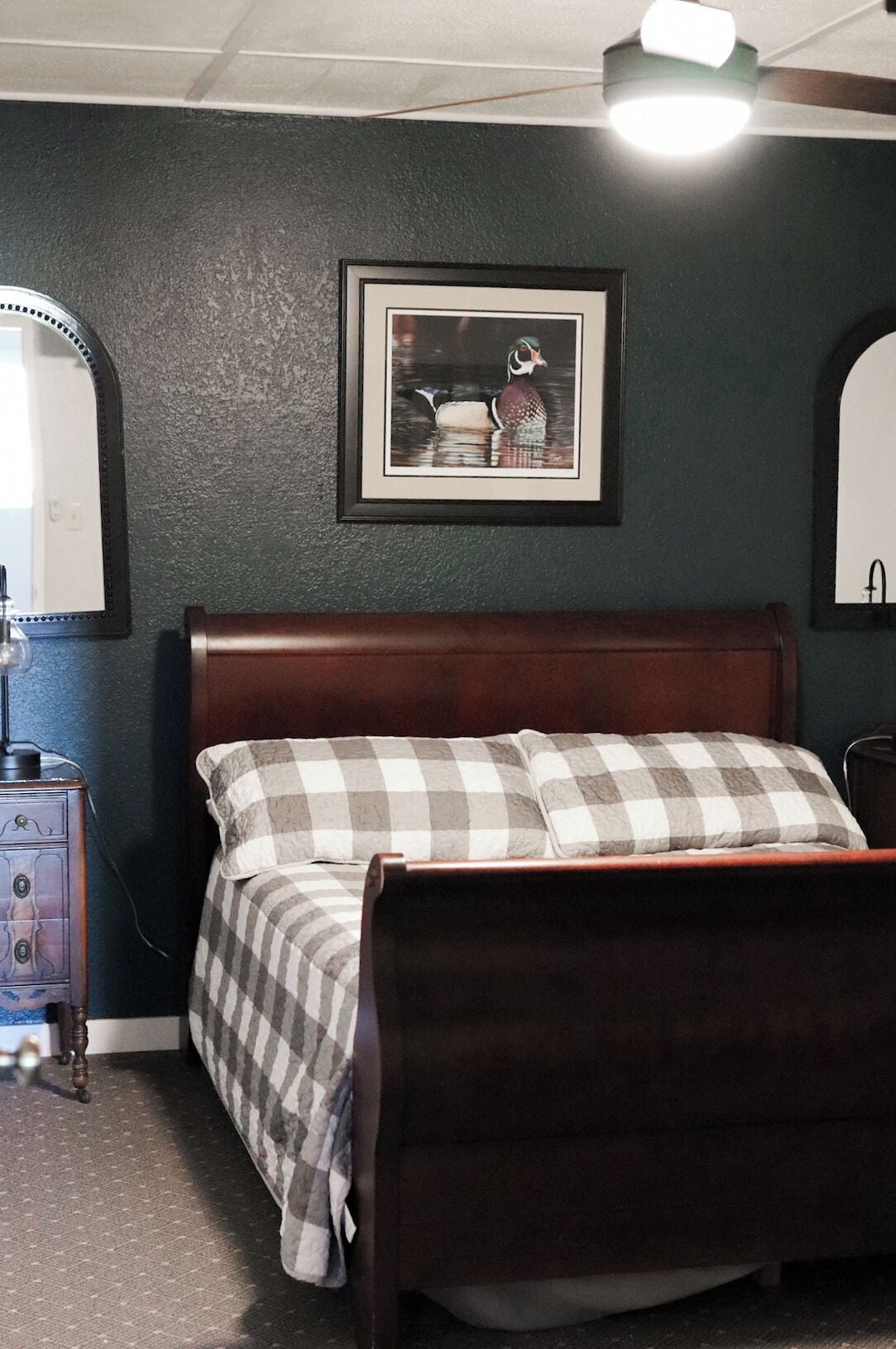 Une chambre à coucher avec un lit en bois, une literie à carreaux, un tableau encadré au mur et une commode avec un miroir.