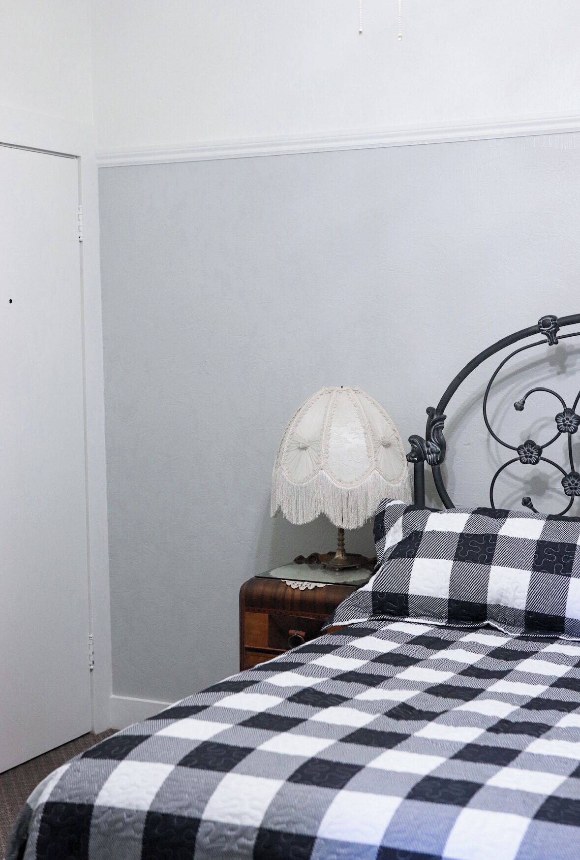 A bedroom with a checkered bedspread, a white lamp on a wooden nightstand, and a metal headboard.