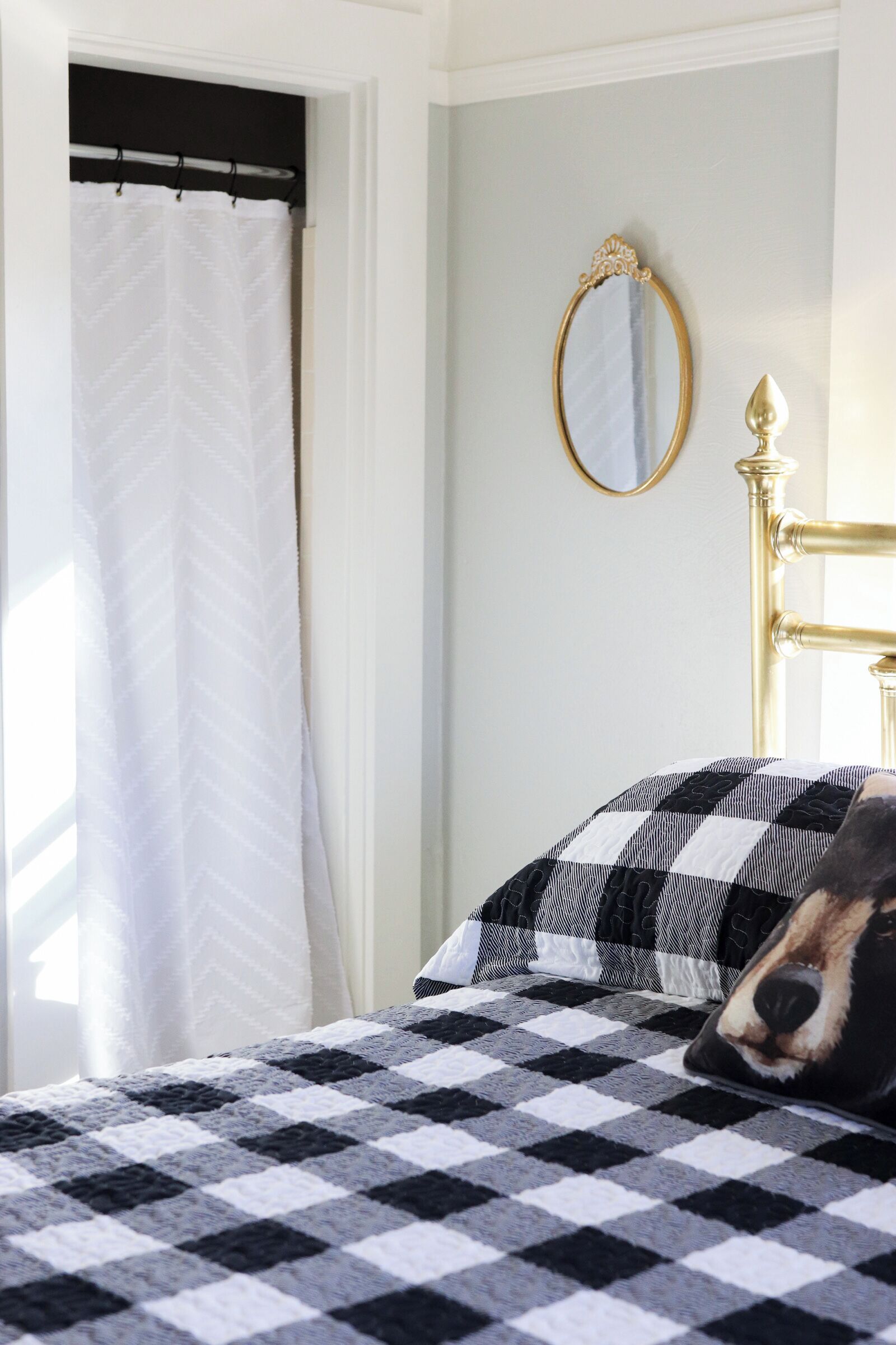 A bedroom with a checkered bedspread, a gold-framed mirror, and a white curtain.