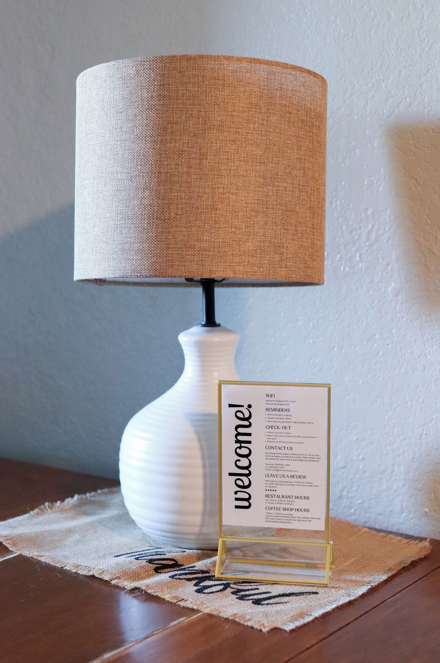 A white table lamp with a beige lampshade, a welcome sign, and a placemat with the word 'welcome' on it.
