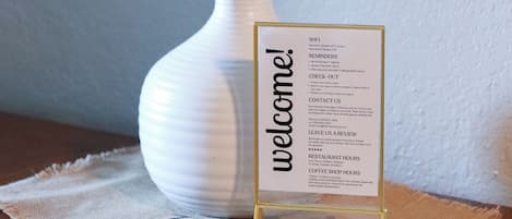 A white table lamp with a beige lampshade, a welcome sign, and a placemat with the word 'welcome' on it.