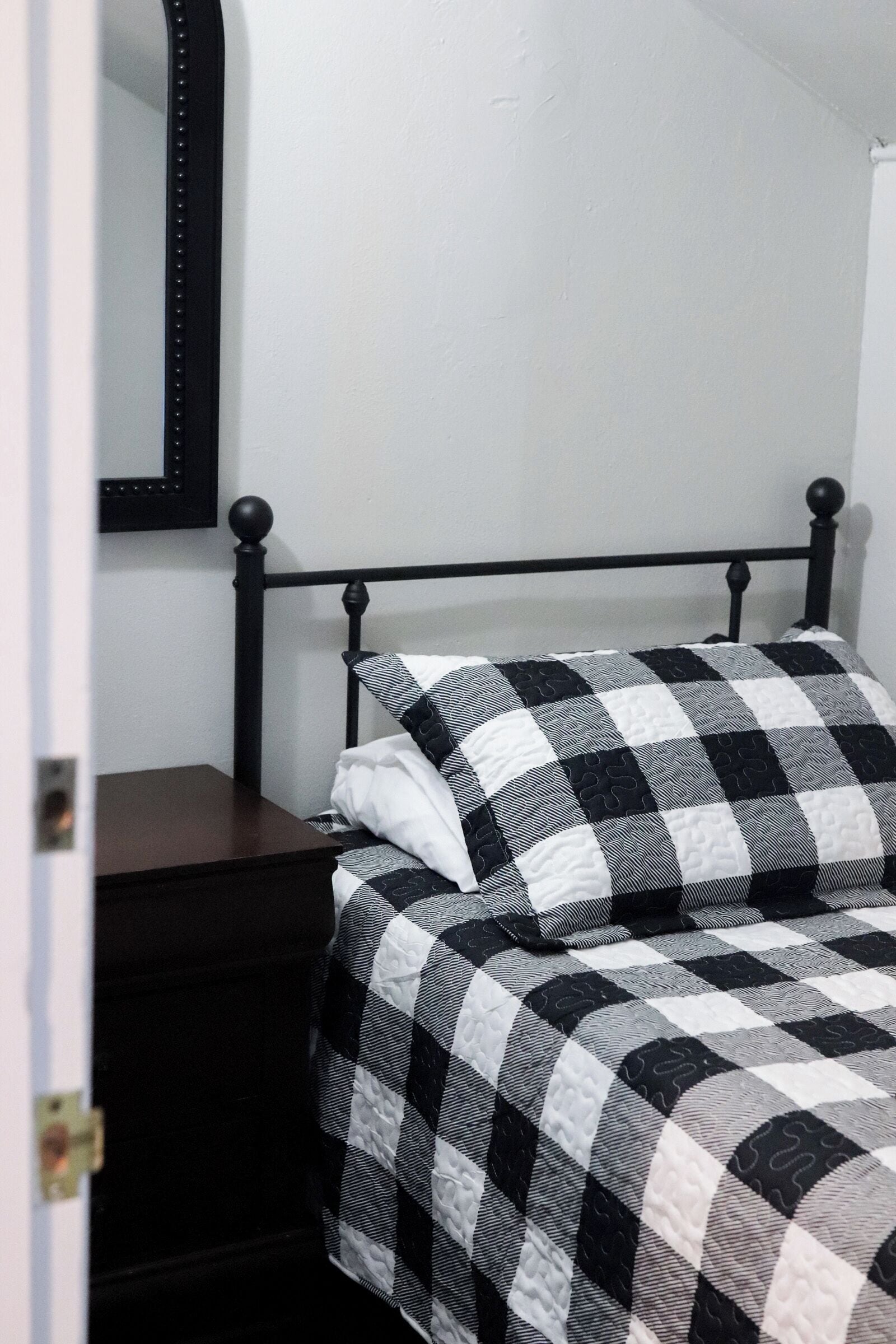 Un lit avec une couvre-lit à carreaux et des oreillers, une table de chevet sombre et un miroir à cadre noir accroché au mur.