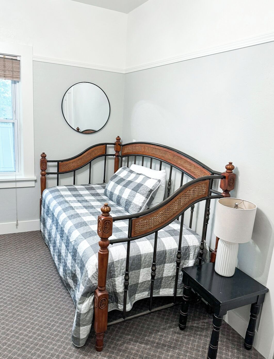 Un lit bien fait, avec une couvre-lit à carreaux et deux oreillers, un miroir rond accroché au mur et une table de chevet avec une lampe.