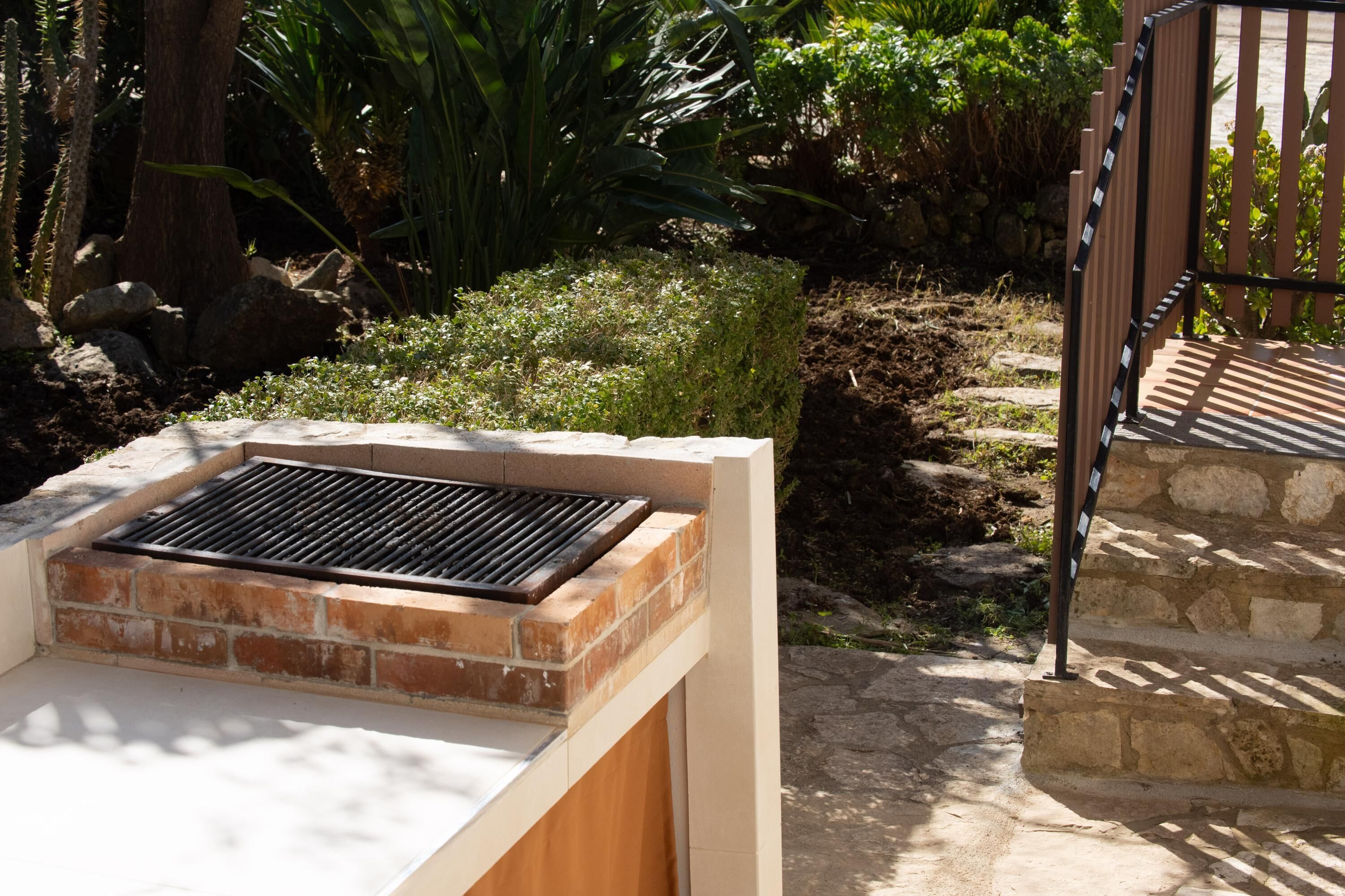 A brick barbecue grill with a metal grate, set against a garden with various plants and a stone staircase.