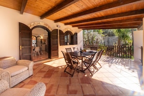 A covered patio with a wooden ceiling, stone arches, a dining table with chairs, and a wicker sofa.