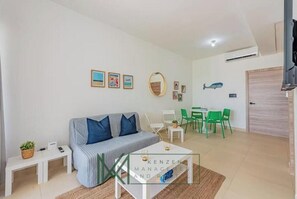 Ein modernes Wohnzimmer mit einer Couch, einem Couchtisch und grünen Sesseln. An den Wänden hängen gerahmte Bilder und auf einem kleinen Tisch steht eine Pflanze.