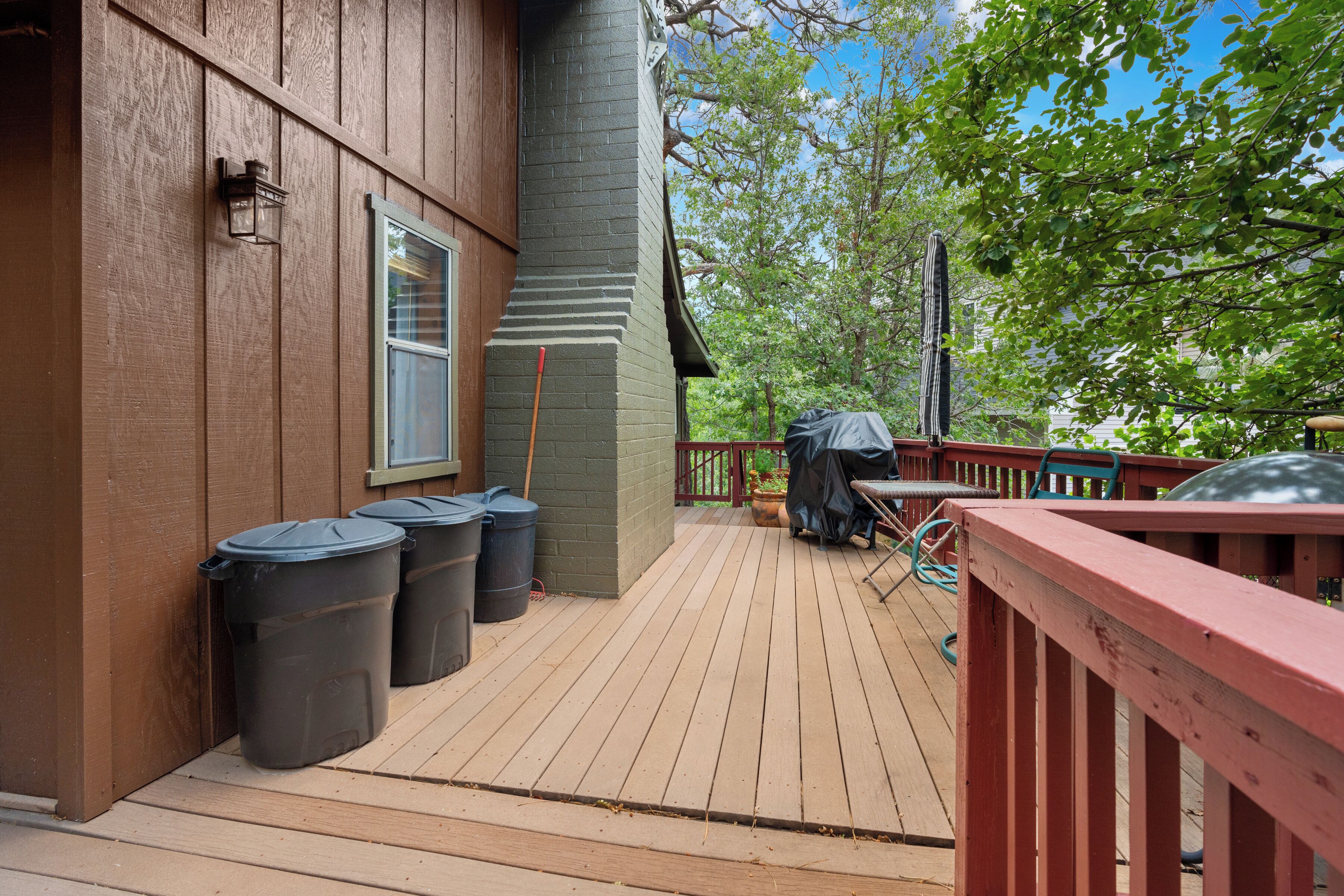 A wooden deck with trash bins, a barbecue grill, and a covered table.