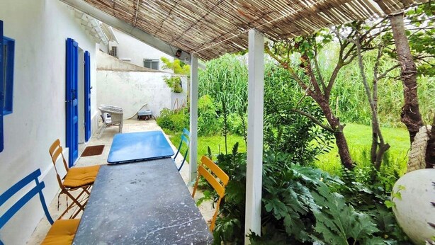 A covered outdoor seating area with a table and chairs, surrounded by greenery and a white wall with blue accents. - La Maisonette Cambon, Charme en Camargue (Saintes-Maries-de-la-Mer)