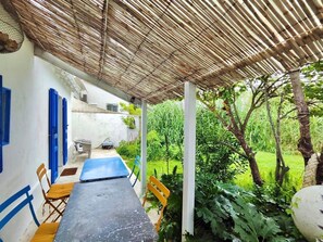 Un área de estar al aire libre cubierta con una mesa y sillas, rodeada de follaje y una pared blanca con detalles azules.