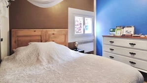 A bedroom with a wooden headboard, a white bedspread, a white dresser with black handles, and a window with floral curtains. - La Maisonette Cambon, Charme en Camargue (Saintes-Maries-de-la-Mer)