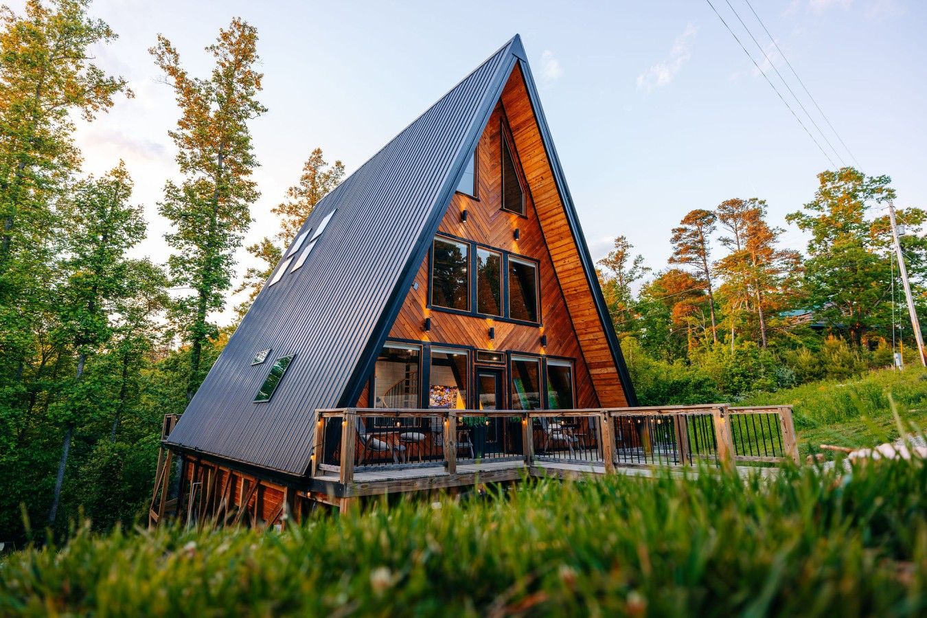 A modern, triangular wooden house with a deck and large windows, surrounded by trees.