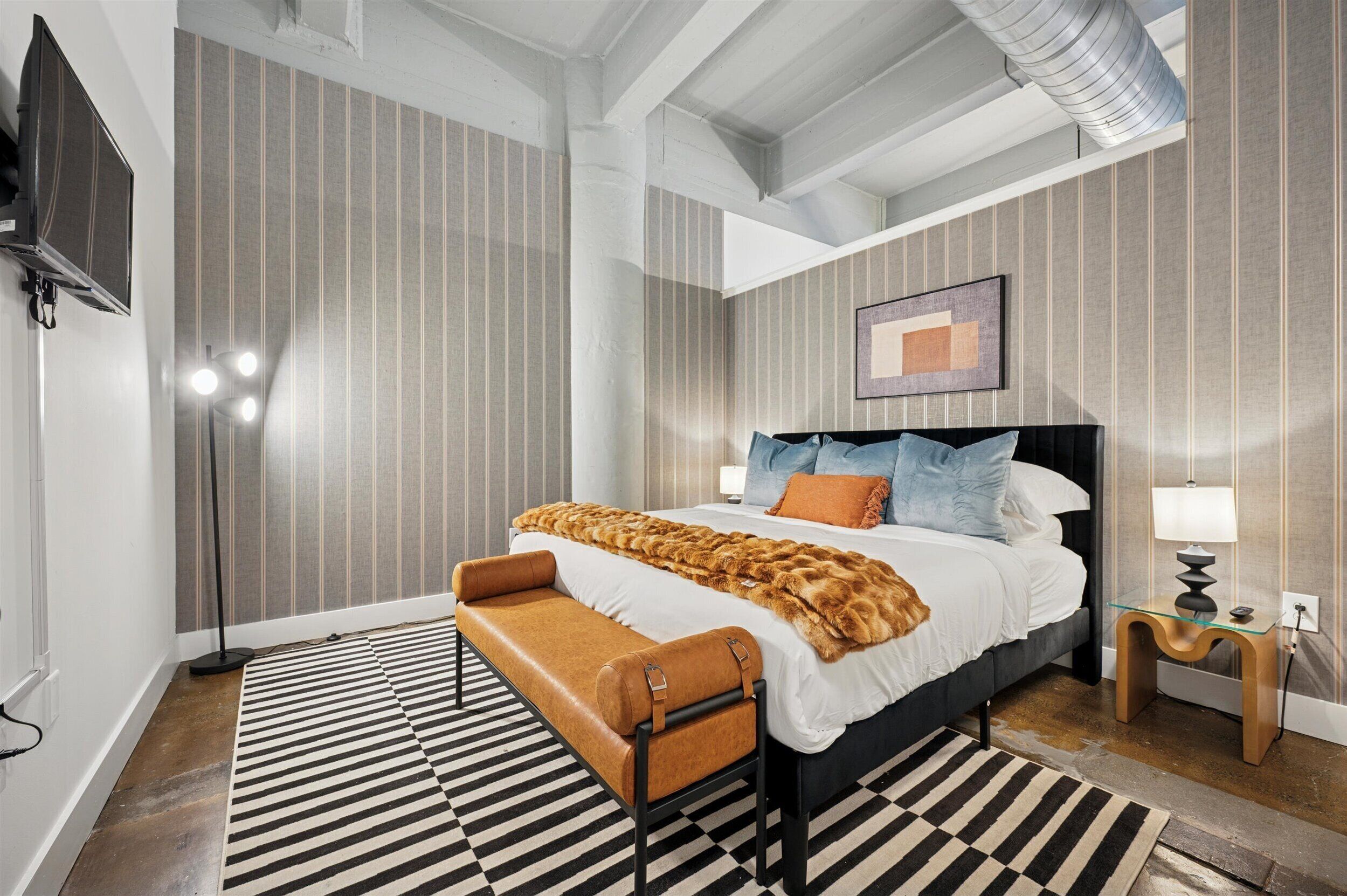 Une chambre à coucher moderne avec un grand lit, un papier peint à rayures, un banc, une télévision et une lampe.