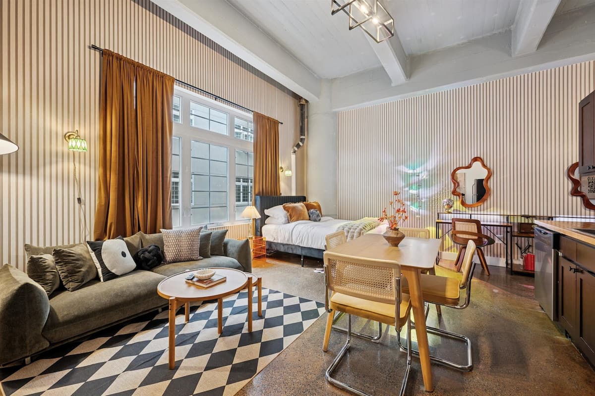 A modern hotel room with a sofa, a dining table, a bed, and a kitchen area.