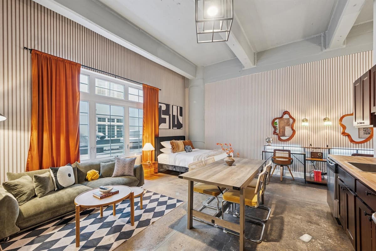A modern hotel room with a large bed, a sofa, a dining table, and a kitchen area.