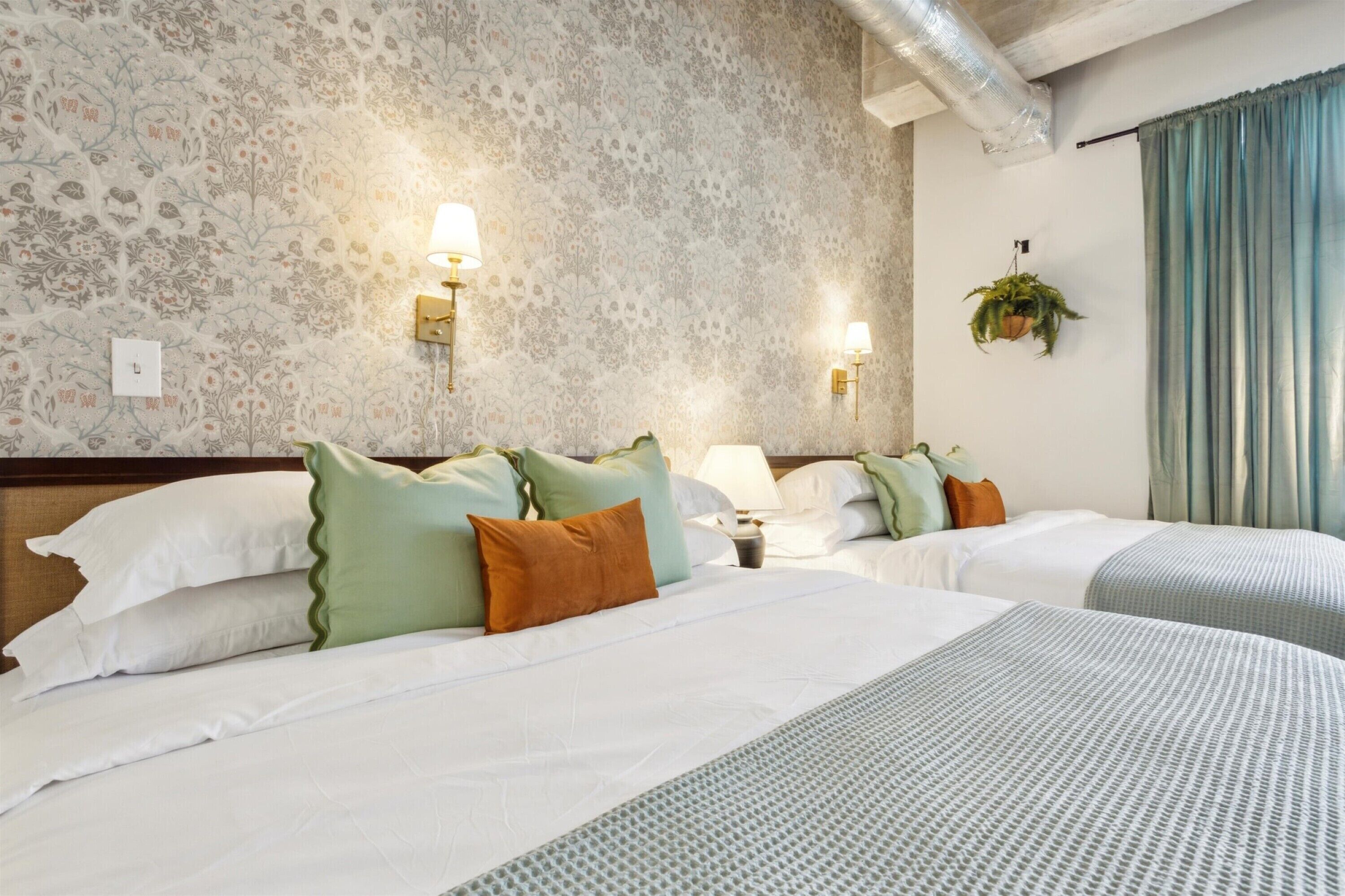 A hotel room with two beds, floral wallpaper, wall sconces, a hanging plant, and a window with curtains.