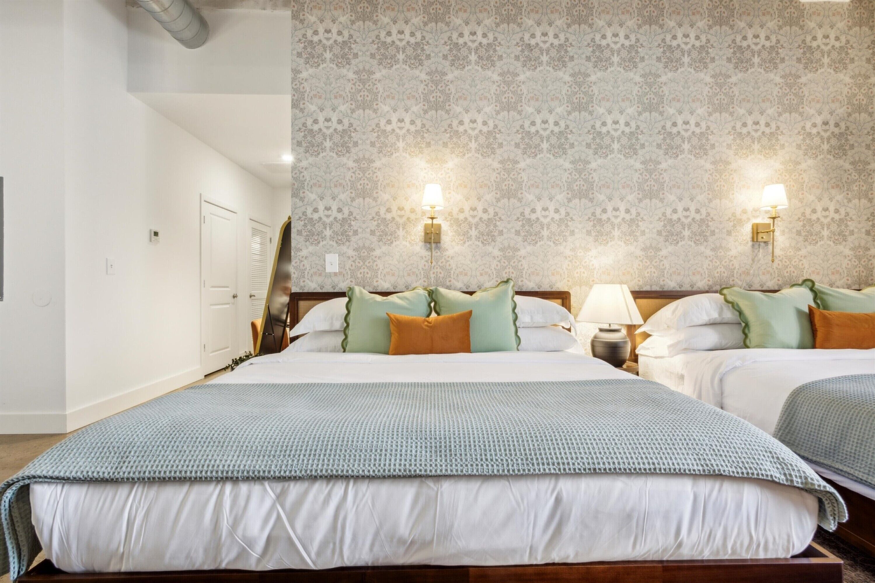A hotel room with two beds, a patterned wallpaper, and wall-mounted lamps.