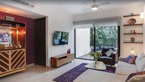 Un salón moderno con sofá, mesa de centro y televisor montado en la pared. Hay una puerta corrediza de vidrio que da a un balcón con vistas a árboles.