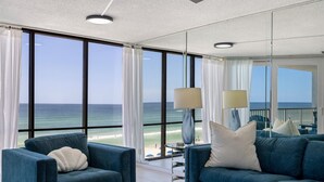 A modern living room with large windows offering a view of the ocean. There are two blue sofas and a glass coffee table. - Seabreeze Bliss @ Edgewater - 5th floor, private balcony, direct beachfront (Panama City Beach)
