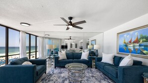 A spacious living room with a large window overlooking the beach, a ceiling fan, and a painting of boats on the wall. - Seabreeze Bliss @ Edgewater - 5th floor, private balcony, direct beachfront (Panama City Beach)