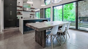 Een moderne keuken met een groot eiland, witte stoelen en grote ramen met uitzicht op groen.