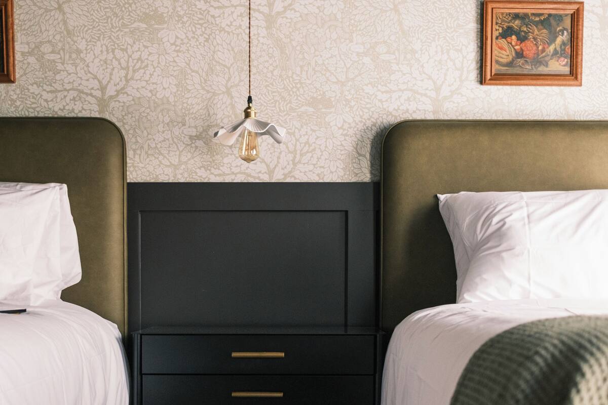 A hotel room with two beds, a nightstand, a hanging light fixture, and a floral-patterned wallpaper.