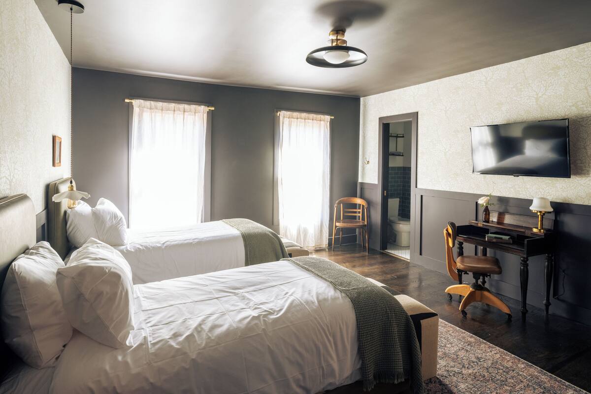 A hotel room with two beds, a desk, a chair, a TV, and a bathroom visible through an open door.