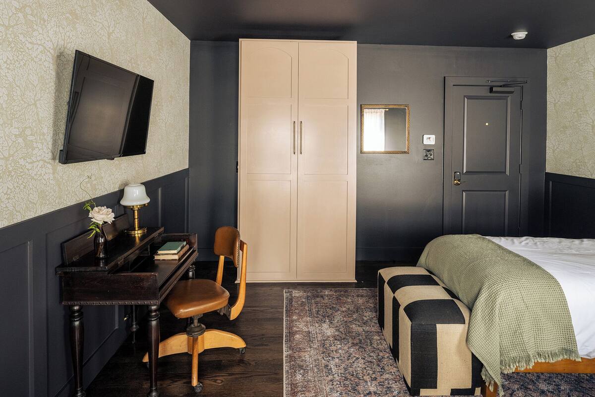 A bedroom with a bed, a desk with a chair, a wardrobe, a television, and a mirror.