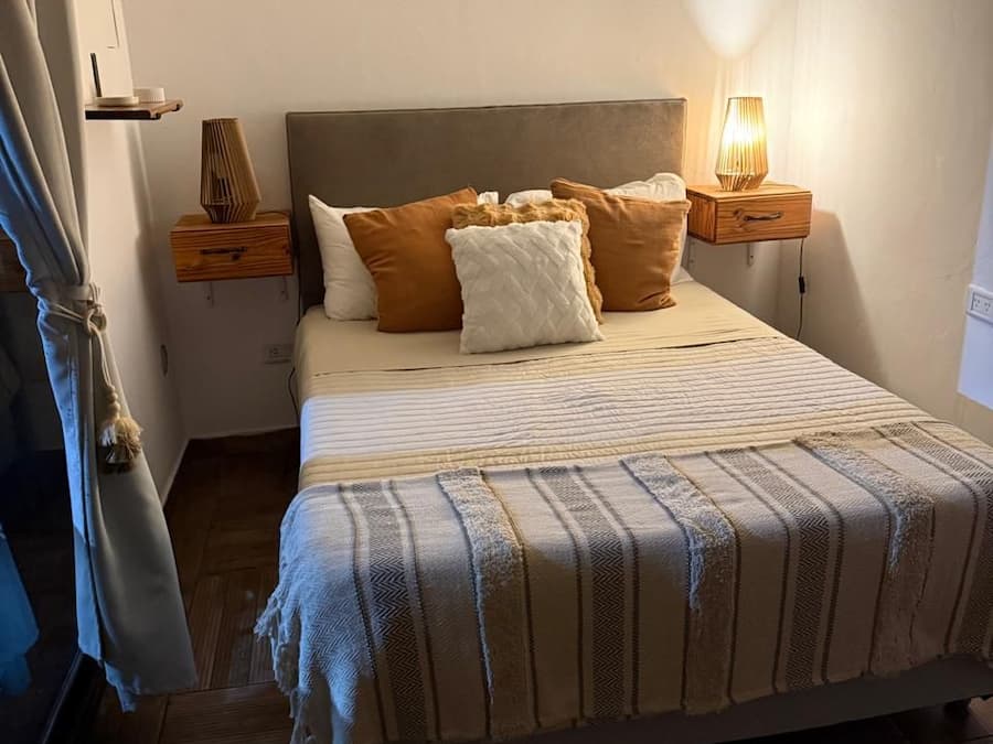 A bedroom with a bed, pillows, a bedside table with a lamp, a wall-mounted artwork, and a wooden floor.