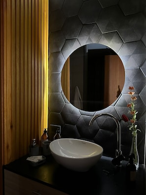 A modern bathroom with a large round mirror, a white sink, and a vase with flowers. - Anas inn (Ezeiza)