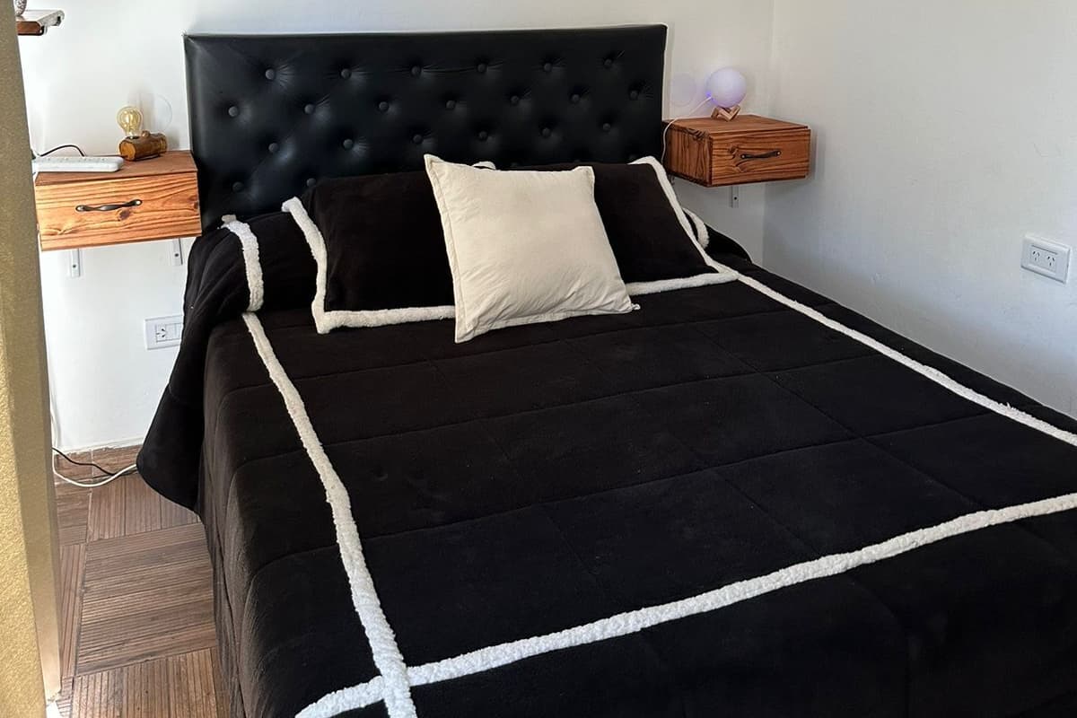 A bedroom with a black and white striped wall art, a tufted black headboard, a black bedspread with white trim, a white pillow, and a wooden nightstand with a lamp.