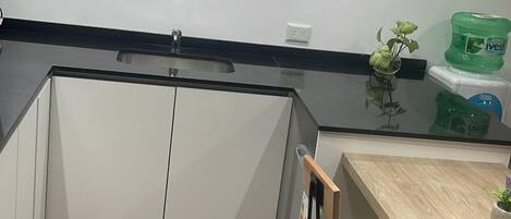 Cocina moderna con armarios blancos, encimera negra y suelo de madera. Hay un taburete de bar y una pequeña mesa con una planta.