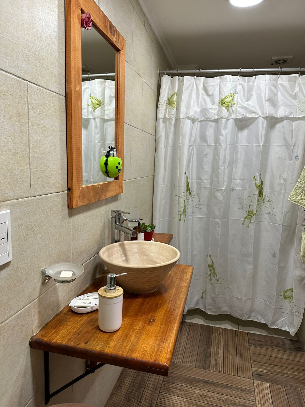 A bathroom with a wooden vanity, a large vessel sink, a mirror, a shower curtain with floral patterns, and a potted plant.