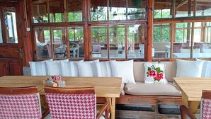 A rustic wooden interior with a large windowed seating area, stone flooring, and houndstooth patterned chairs. - Dilara Huzur Motel (Ayvacik)