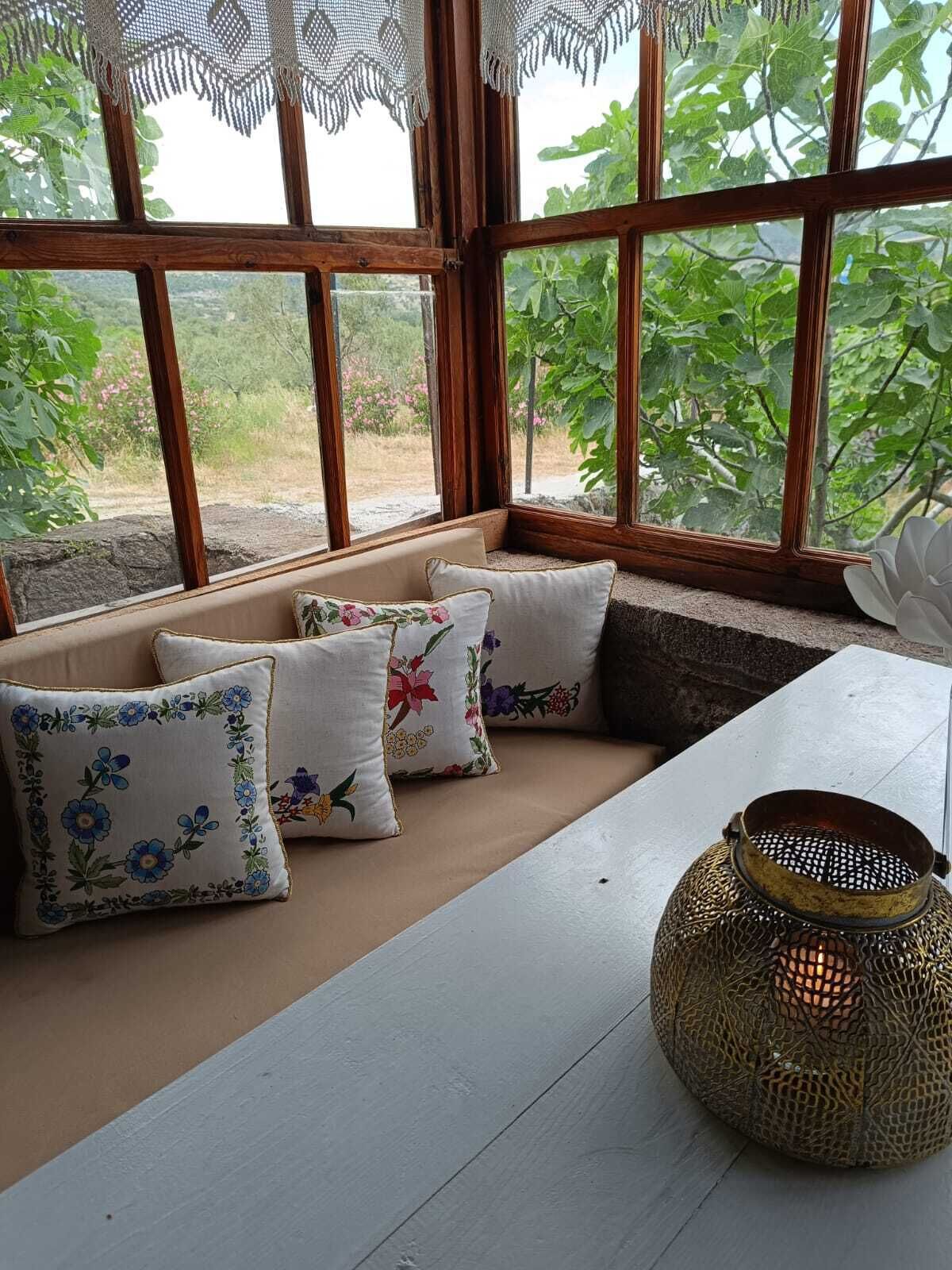 Un rincón acogedor con bancos acolchados y cojines de estampado floral, un farol tejido y ventanas amplias con vistas al verdor.
