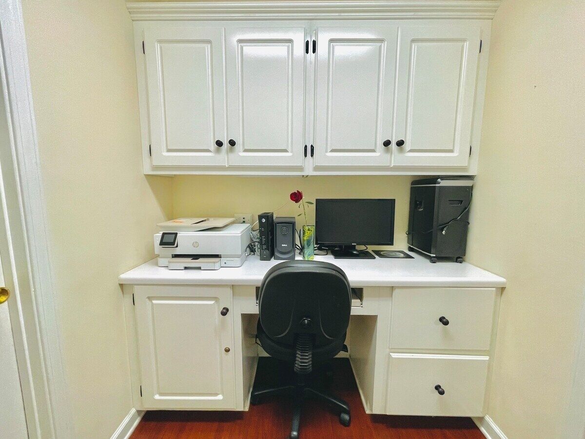 A white desk with a printer, monitor, and computer tower. There is a black office chair in front of the desk.