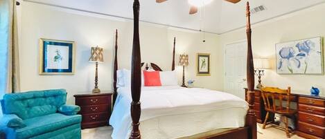 A four-poster bed with a ceiling fan, a teal armchair, a wooden desk, and a mirror.