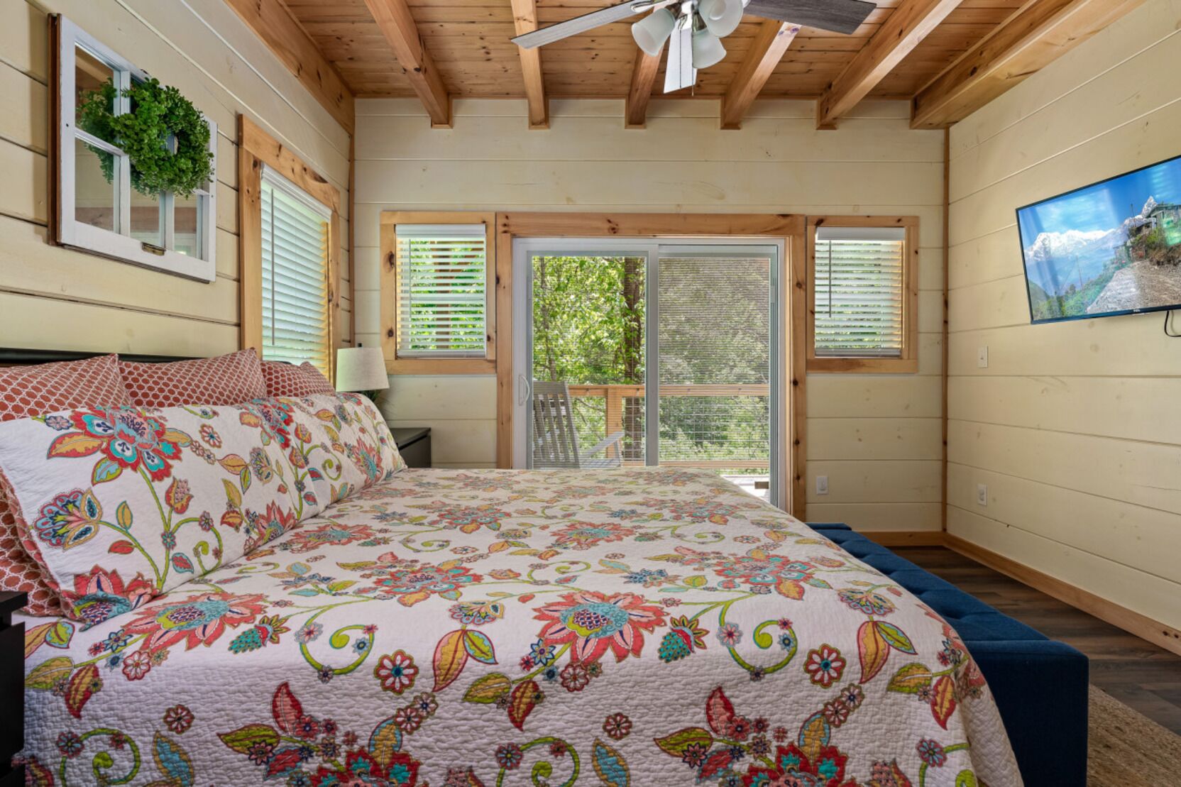 A bedroom with a large bed, wooden ceiling beams, a window with a wreath, and a view of greenery outside.