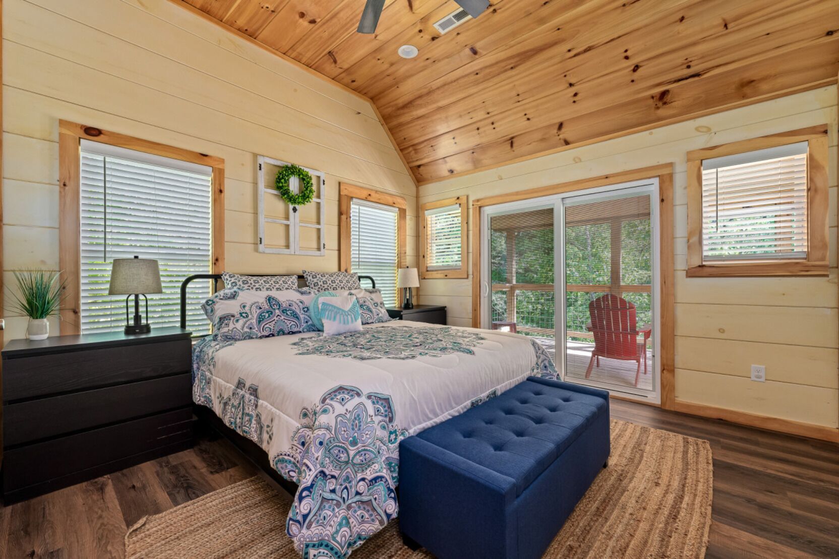 A bedroom with a large bed, a nightstand, a dresser, a window with blinds, a door, and a view of the outdoors.