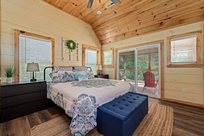 A bedroom with a large bed, a nightstand, a dresser, a window with blinds, a door, and a view of the outdoors.