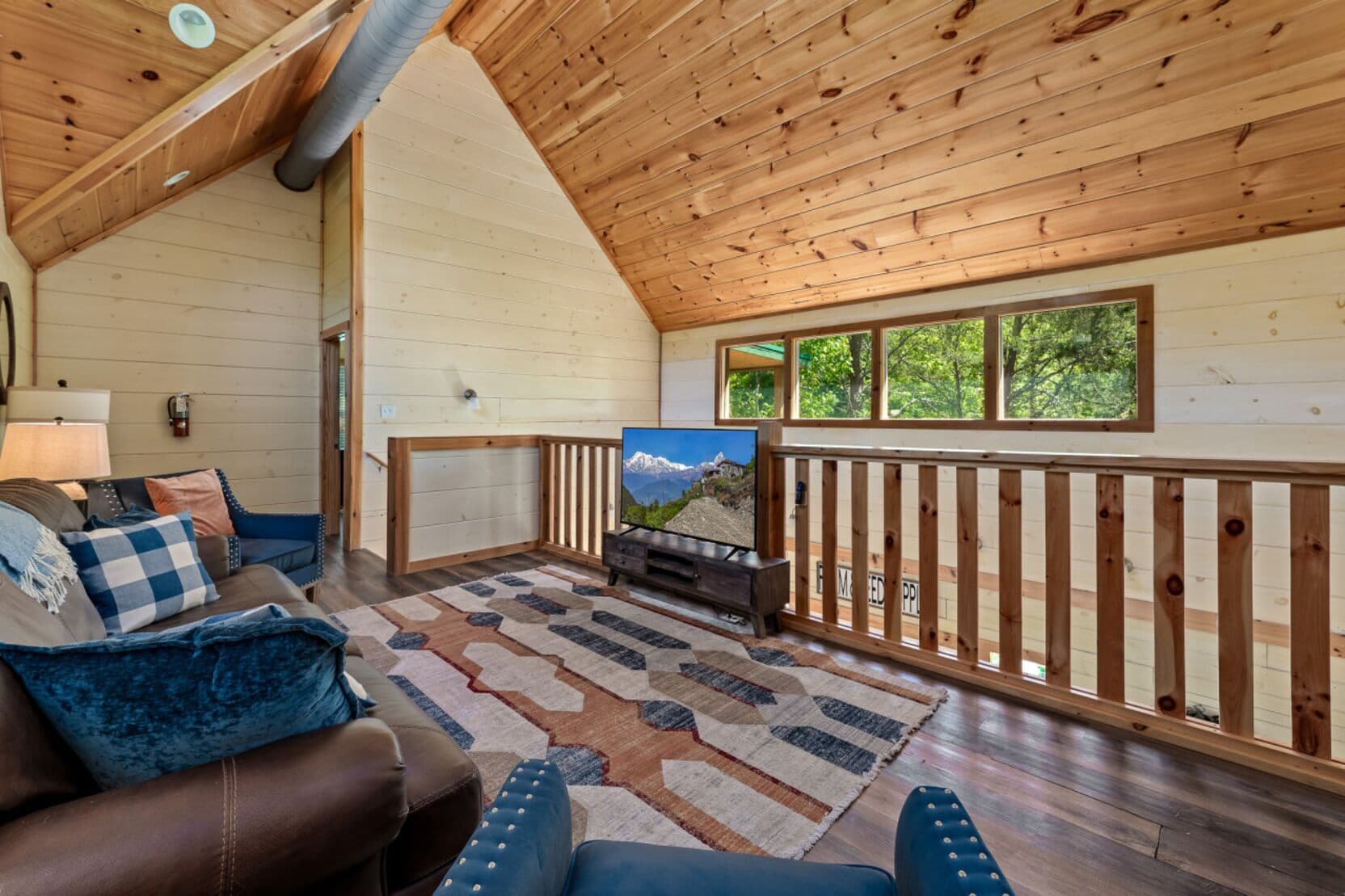 A cozy cabin interior with a wooden ceiling, a large window overlooking greenery, a fireplace, and a comfortable seating area with a patterned rug.